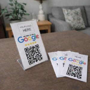 A lifestyle product photograph of custom Google review marketing tools arranged on a brown wooden tabletop. On the left, a vertical clear acrylic sign holder stands upright, displaying a printed template with a placeholder reading 'YOUR LOGO HERE', the multi-colored 'REVIEW US ON Google' graphic, five yellow stars, and a prominent square QR code. To the right, a fanned-out stack of three identical printed paper cards lies flat on the table, with a blurred living room couch and houseplant visible in the background.