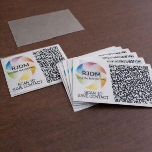 A high-angle studio photograph of custom print branding assets arranged on a dark wood texture. On the left sits a single square card enclosed in a clear protective plastic sleeve, and to the right is a fanned-out stack of identical square cards. Each card displays the multi-colored circular RJDM Digital Marketing logo, a large complex QR code, and the instructional text 'SCAN TO SAVE CONTACT'. A translucent rectangular piece of backing material lies flat in the upper background.