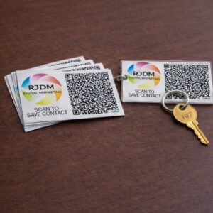 A high-angle product photograph displaying printed marketing assets on a dark wooden surface. On the left is a stacked bundle of square cards featuring the colorful, circular RJDM Digital Marketing logo, a prominent square QR code, and the text 'SCAN TO SAVE CONTACT'. To the right sits a matching rectangular clear acrylic key ring housing the same brand design, attached to a brass key stamped with the number 107.